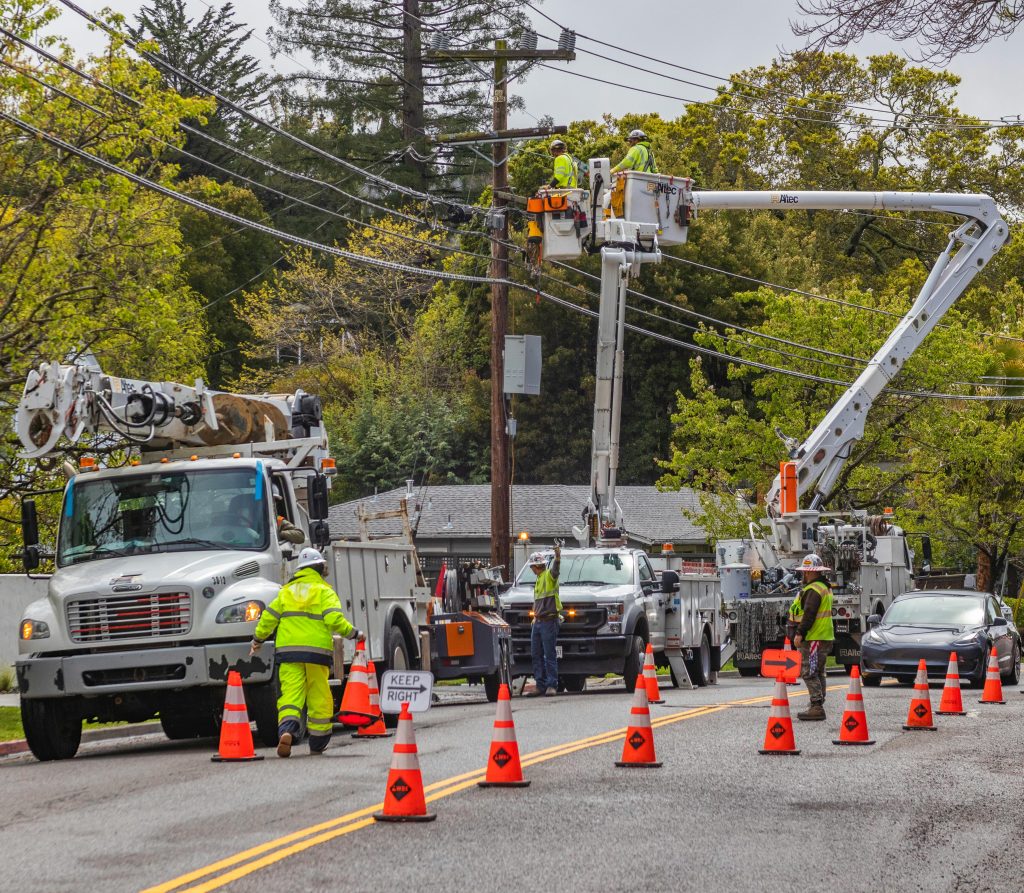 What We Believe 3 Utility workers in reflective gear repair power lines using bucket trucks and cones on a suburban street.